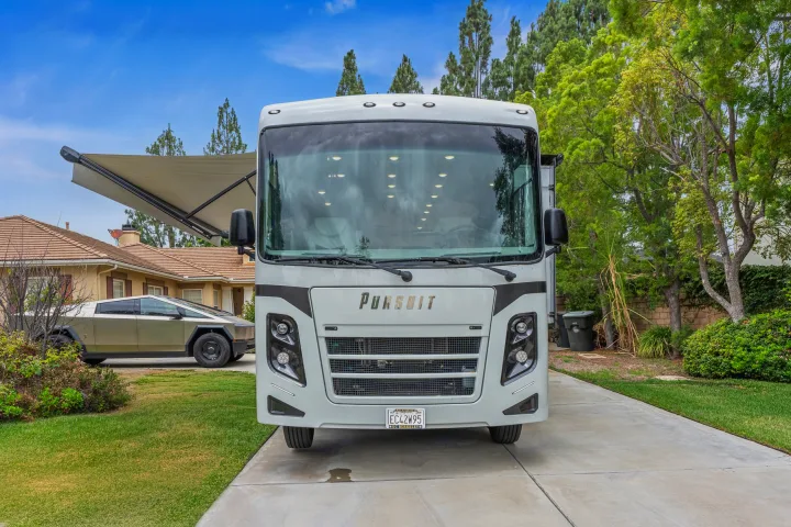 White RV rental with awning extended on a driveway, a Cybertruck, house, and green trees in the background.. Coachmen Pursuit 2024