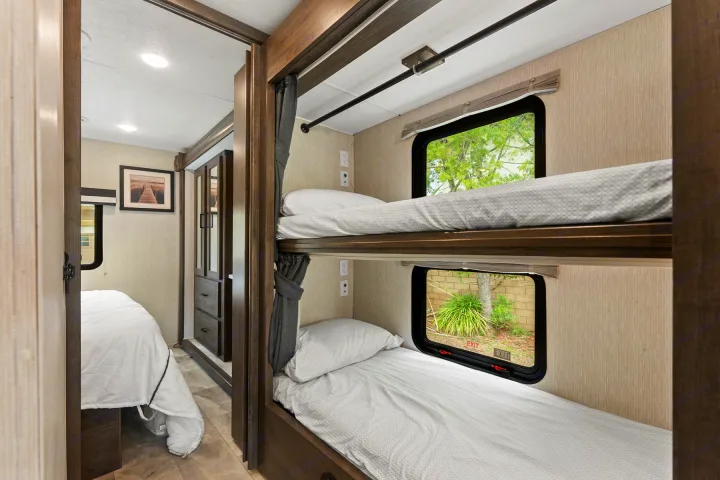 Interior of an RV rental showing modern bunk beds with windows, alongside a glimpse into an adjacent private bedroom with a larger bed.. Coachmen Pursuit 2024