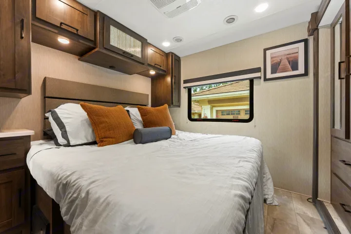 Inviting RV rental bedroom with a comfy bed, overhead cabinets, and a window looking out to a home.. Coachmen Pursuit 2024