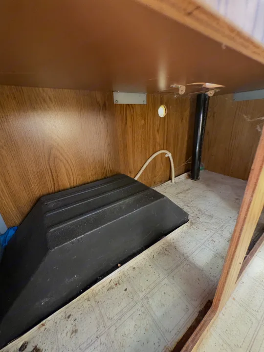 Under-cabinet view in an RV rental, showing a large black cover over utilities on a tiled floor, with pipes and hoses along wood-paneled walls.. Coleman Destiny Roanoke 1991
