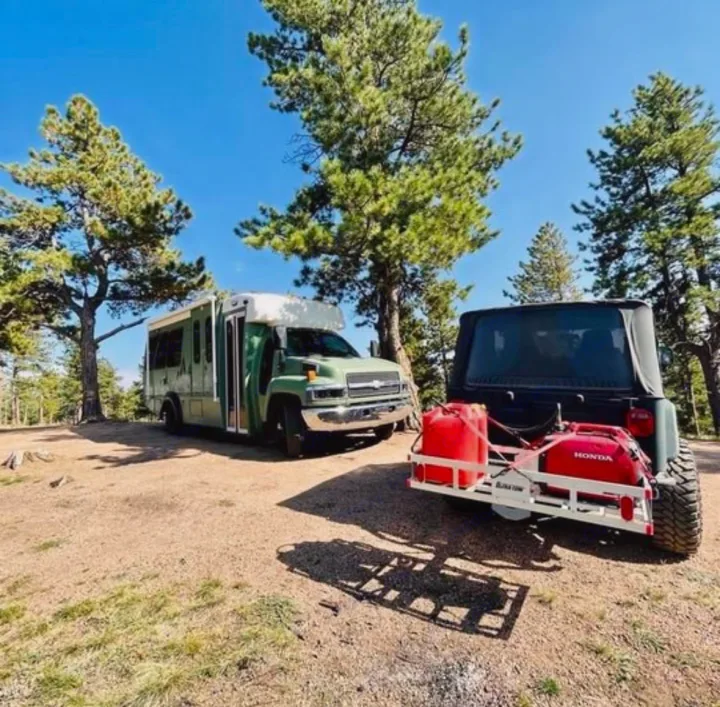 Green converted bus RV rental and a Jeep with red fuel cans, parked among pine trees on a sunny day.. Chevy C4500 Kodiak 2008