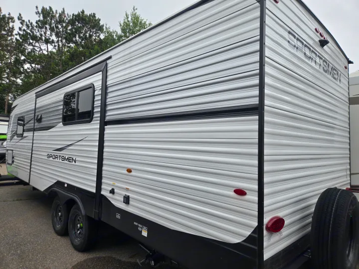 This white Sportsmen travel trailer, ideal for RV rental, offers adventure. Side view shows tires, hookups, and a rear spare.. KZ Sportsmen 291BHK 2027