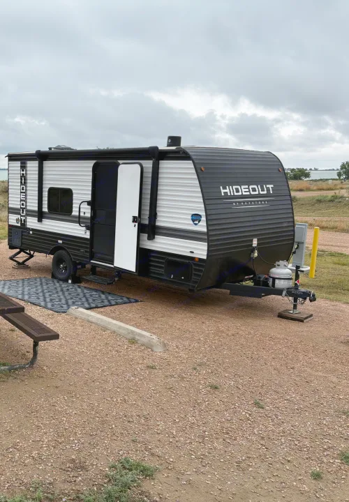 This 'Hideout by Keystone' rv rental is set up at a lakeside campsite under a grey sky. Perfect for an outdoor adventure.. Keystone RV Hideout Sport 2024