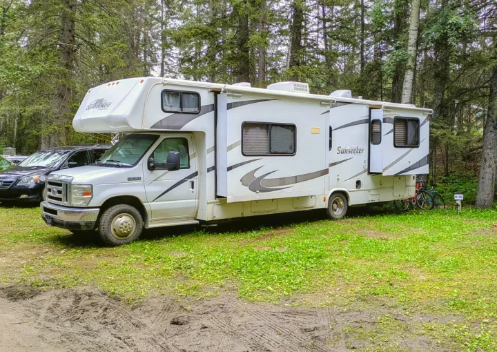 This white Sunseeker rv rental with a slide-out is at a muddy forest campsite (R 35). A car and bike are visible.. Forest River Sunseeker 2010