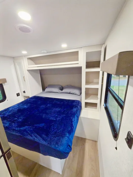 Cozy RV rental bedroom features a comfortable bed with a blue blanket, overhead storage, and a window. Perfect for travel.. Grand Design RV Transcend Xplor 26BHX 2027