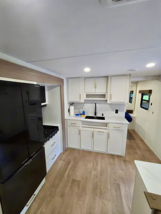 Modern RV rental kitchen features white cabinets, black appliances, light wood flooring, and gold hardware.. Grand Design RV Transcend Xplor 26BHX 2027