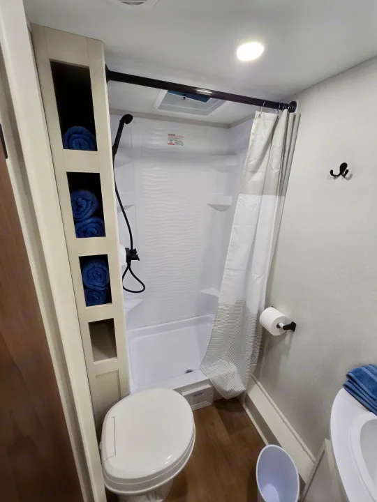 A compact RV rental bathroom featuring a shower, toilet, sink, and organized shelves with blue towels.. Grand Design RV Transcend Xplor 26BHX 2027