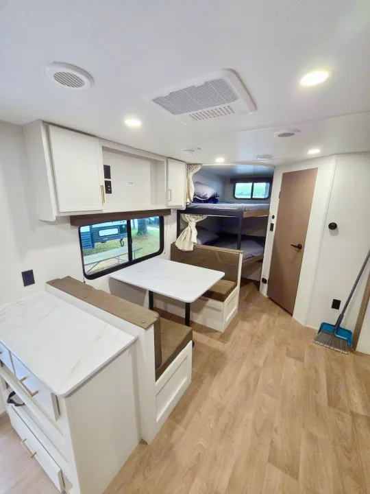 Bright RV rental interior showing a modern dining booth, kitchen counter, and bunk beds. Light wood floors and white cabinetry.. Grand Design RV Transcend Xplor 26BHX 2027
