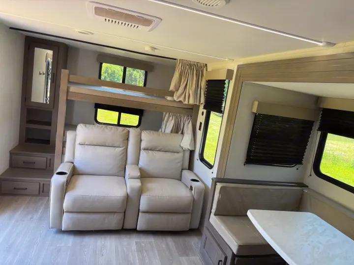 Light-toned rv rental interior featuring bunk beds, two recliners, and a dinette area. Bright windows show green outdoors.. Grand Design Transcend 2025