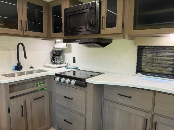 Modern RV rental kitchen featuring a sink, microwave, stovetop, coffee maker, and light wood cabinets. Ready for your travels!. Grand Design Transcend 2025