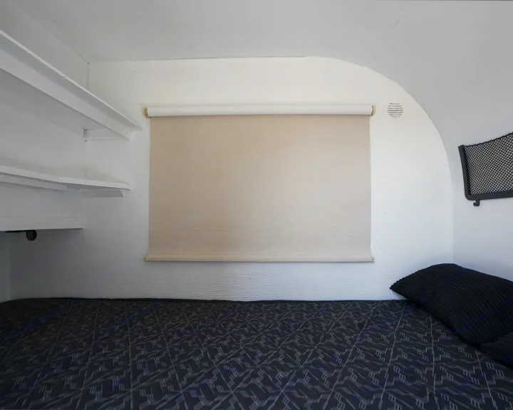 Cozy RV rental sleeping area featuring a dark patterned bed, pull-down blind, built-in shelves, and wall vent.. Micro-Lite Micro Camper 1987