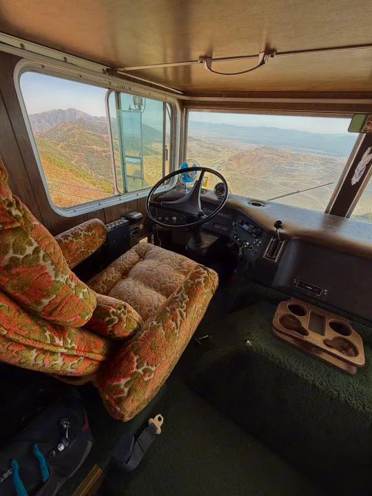 Inside a vintage rv rental: floral seats, shag carpet, and a steering wheel frame a panoramic mountain view.. International 510A 1973