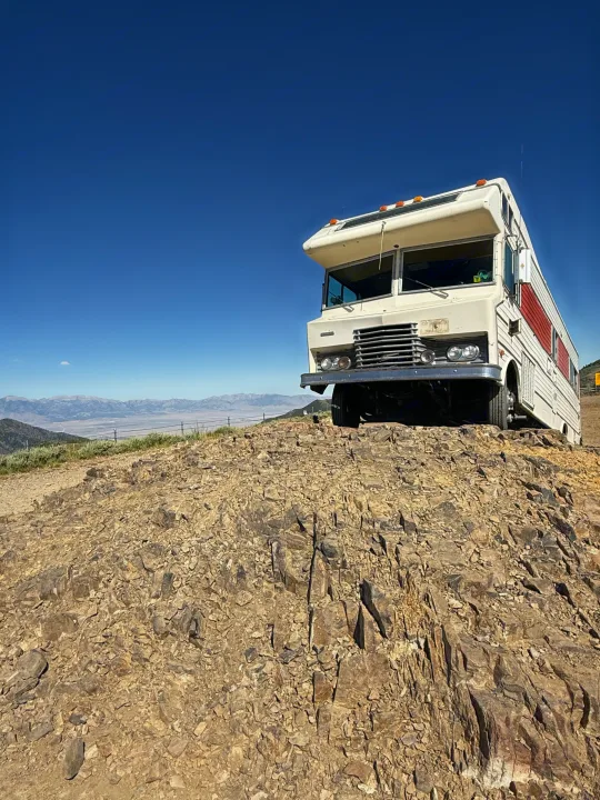 Experience unique journeys! A vintage RV rental sits high on a rocky ridge, showcasing vast mountain panoramas under a clear blue sky.. International 510A 1973