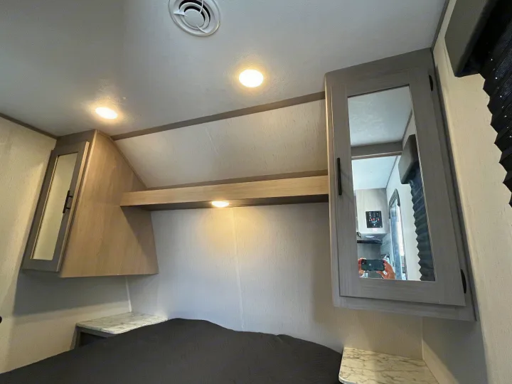 Inside an RV rental, a bedroom features overhead lighting, storage cabinets, and a comfortable bed. Photographer visible in mirror.. Dutchmen Coleman Lantern LT 2022