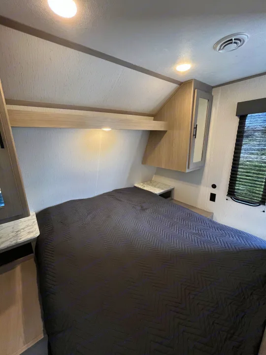 RV rental bedroom: a modern space with a dark gray quilted bed, overhead cabinets, small side tables, and a window with blinds.. Dutchmen Coleman Lantern LT 2022