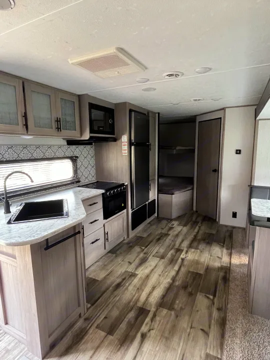 Bright rv rental interior: modern kitchen with light wood cabinets, white counters, wood-look floor, and a glimpse of bunk beds.. Dutchmen Coleman Lantern LT 2022