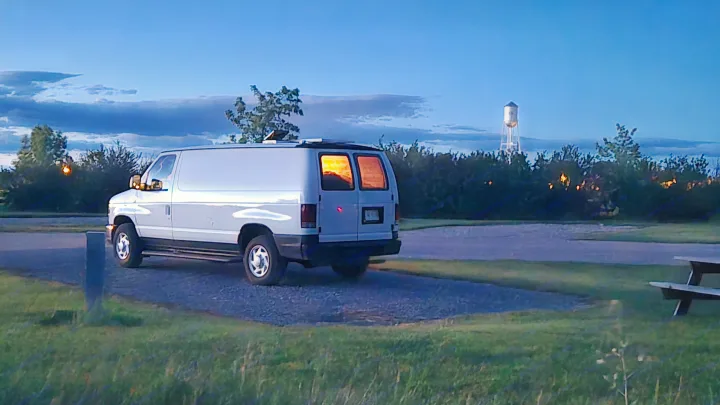RV rental camper van with solar panels, glowing rear windows, parked at dusk. A distant water tower adds to the tranquil scene.. Ford Econoline 2011