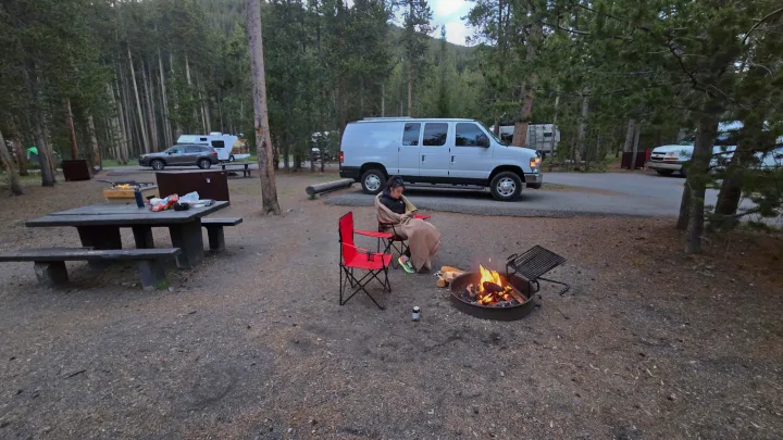 Wooded campsite scene: A person by a warm campfire near a white RV rental van, picnic table, and red chairs.. Ford Econoline 2011