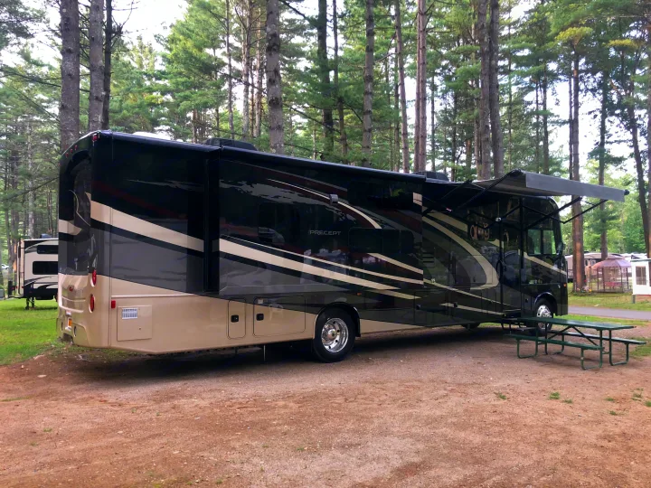 A large, striped Class A rv rental is parked in a serene, pine-filled campground. Its awning is extended, with a green picnic table nearby.. Jayco Precept 2019