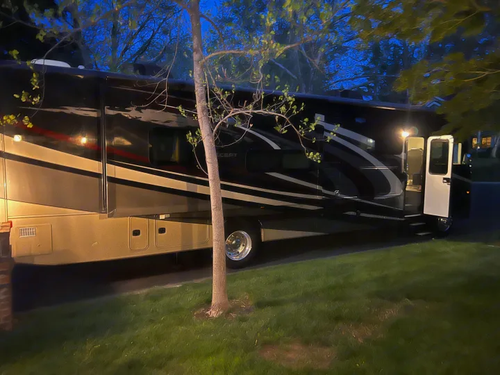 Luxury RV rental parked at night, with its door open and steps illuminated. A slender tree stands in front of the motorhome.. Jayco Precept 2019