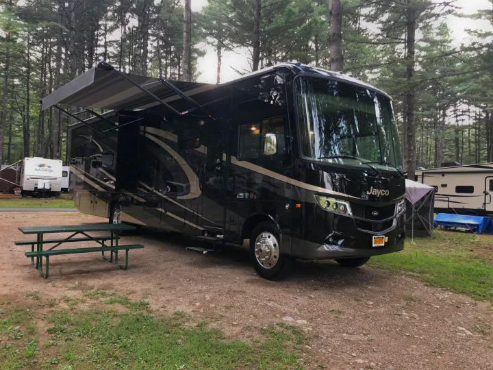 Black Jayco motorhome RV rental at a wooded campsite with picnic table and awning extended.. Jayco Precept 2019