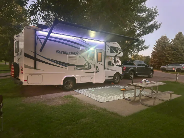 Spacious Sunseeker RV rental at a serene campsite with a lit awning, outdoor rug, picnic table & truck.. Forest River Sunseeker LE 2018