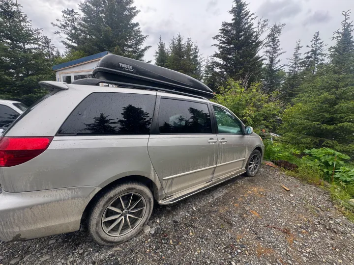 Dirty silver minivan with Thule cargo box, parked by trees on gravel. Perfect for a journey like an rv rental trip.. Toyota Toyota Sienna XLE 2005