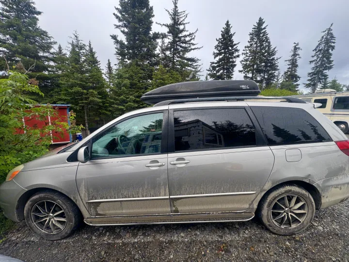 Dirty minivan with roof cargo box parked on gravel near an RV rental, surrounded by tall evergreen trees.. Toyota Toyota Sienna XLE 2005