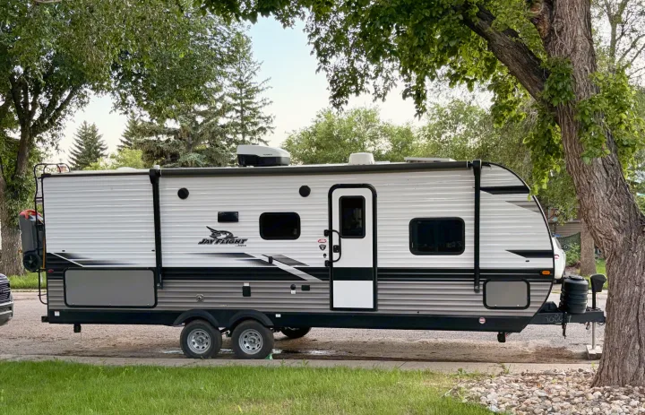 White and grey Jayco Jay Flight RV rental travel trailer, ready for your next camping or road trip, parked under green trees.. Jayco Jay Flight 2023