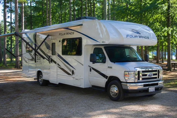 White Four Winds Class C RV rental with an extended awning, parked on gravel amidst trees. Ideal for camping near water.. Thor Four Winds 2024