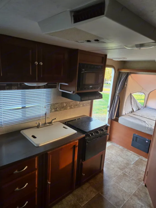 Functional kitchen in an rv rental, featuring a sink, stove, and microwave. An open pop-out bed area is visible.. Dutchmen Kodiak 2010