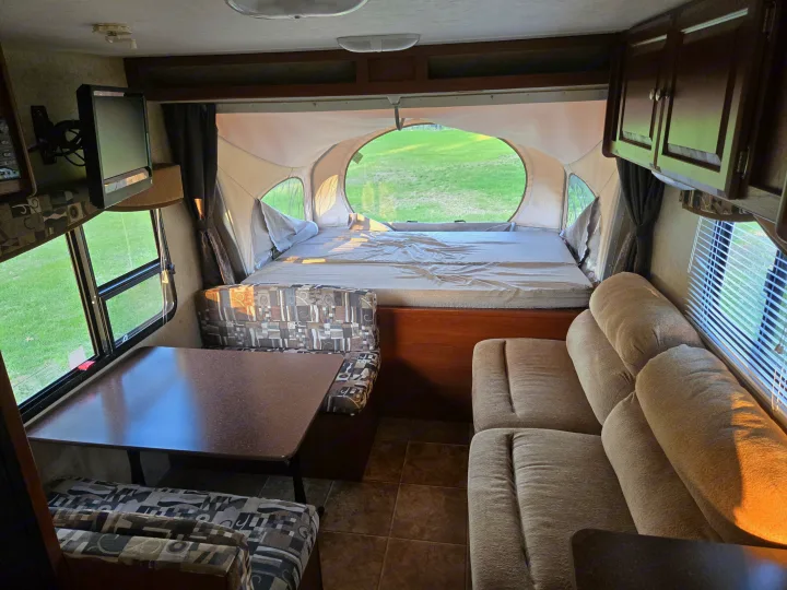 View inside an RV rental showing a patterned dinette, plush sofa, and a large pop-out bed overlooking a green field.. Dutchmen Kodiak 2010