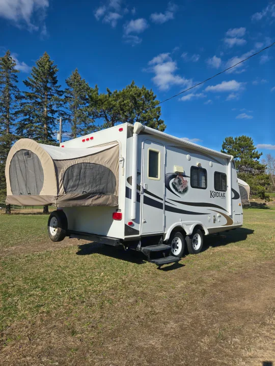 A KODIAK travel trailer RV rental, grey and white with a canvas pop-out tent, parked on grass under a blue sky.. Dutchmen Kodiak 2010