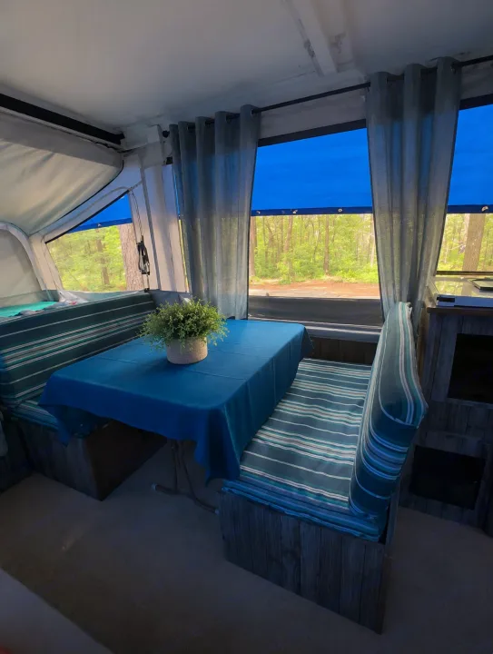 Inside a vibrant RV rental: a blue table, striped bench seating, and large windows overlooking a serene forest.. Coleman Santa Fe 2003