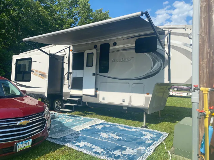 Fifth-wheel RV rental at a sunny campsite with an extended awning, a red SUV, and outdoor rugs on the grass.. Keystone RV Montana High Country 2013