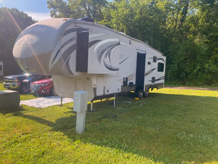 Large fifth-wheel rv rental set up at a sunny campsite, featuring utility hookups, parked vehicles, and lush green trees.. Keystone RV Montana High Country 2013