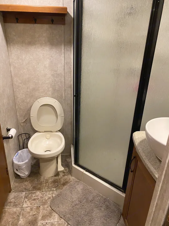 Functional RV rental bathroom with a toilet, frosted shower, and vanity. Small trash can and shelf with hooks.. Keystone RV Montana High Country 2013