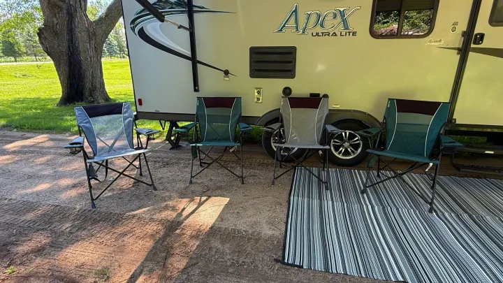 A cozy rv rental campsite with four folding chairs and a striped mat under a large tree, ready for relaxation.. Coachmen Apex 2016