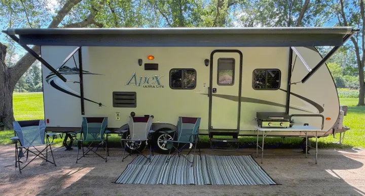 This RV rental campsite setup features an extended awning, chairs, rug, and an outdoor cooking station.. Coachmen Apex 2016
