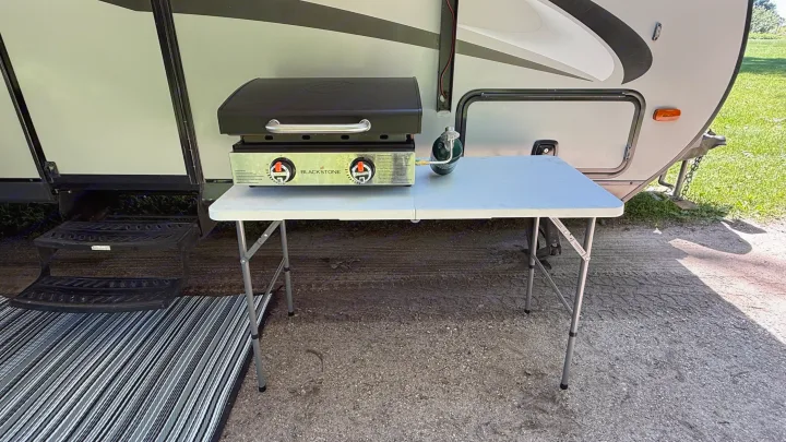**RV rental cooking setup:** Blackstone griddle on a white portable table with propane, beside a light gray RV. Ideal for outdoor meals.. Coachmen Apex 2016