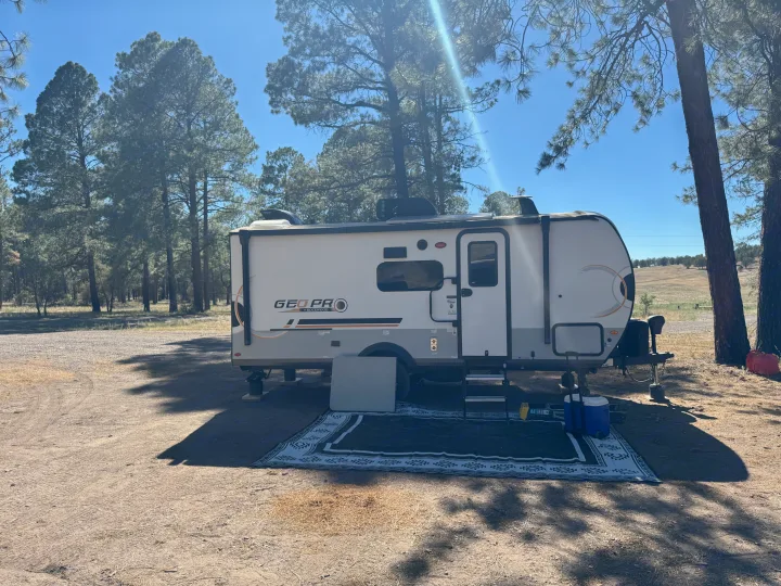 An rv rental is parked and set up for camping under tall pine trees on a sunny day, with an outdoor mat and cooler.. Forest River Rockwood Geo Pro 2022