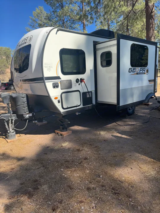White 'GEO PRO' travel trailer (RV rental) at a pine forest campsite. Slide-out extended, power connected on dirt ground.. Forest River Rockwood Geo Pro 2022