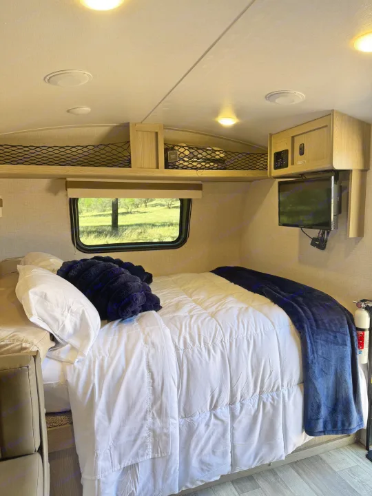 Inside a cozy rv rental bedroom: bed with blue blanket, scenic window, overhead storage, TV, and fire extinguisher.. Forest River Rockwood Geo Pro 2022