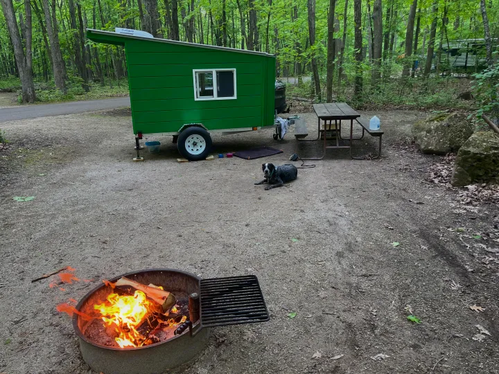 A green rv rental camper is set up at a gravel campsite with a dog, picnic table, and a warm campfire in the woods.. Custom Custom 2025