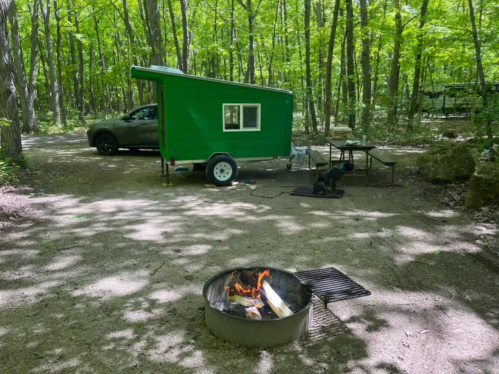 This compact green RV rental provides a cozy camp experience, with a dog resting, a picnic table, and a warm campfire.. Custom Custom 2025