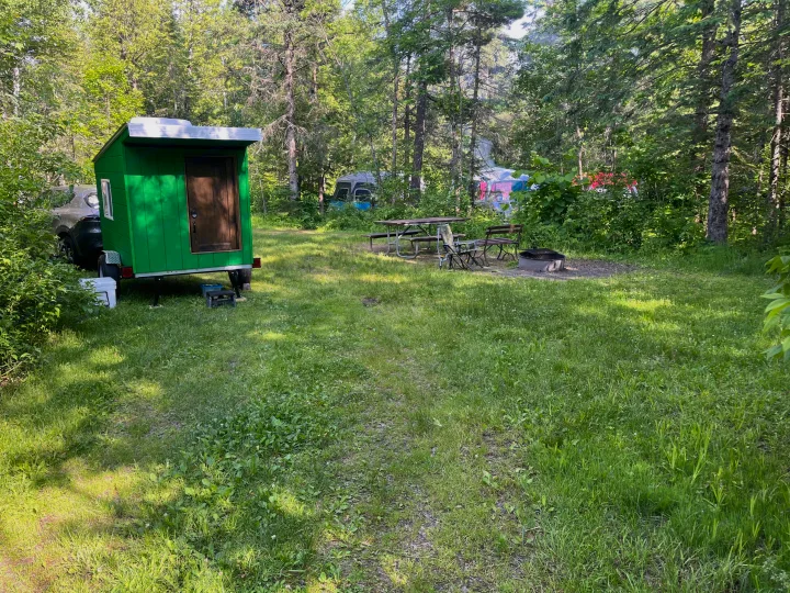 Green RV rental trailer at a sunny campground with a picnic table, chairs, and fire pit among trees.. Custom Custom 2025