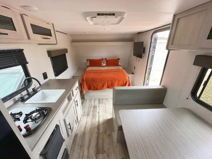 A bright, modern rv rental interior with a kitchen featuring a stove and sink, a cozy bed with orange bedding, and a dining booth.. Crossroads RV Zinger Lite 2026