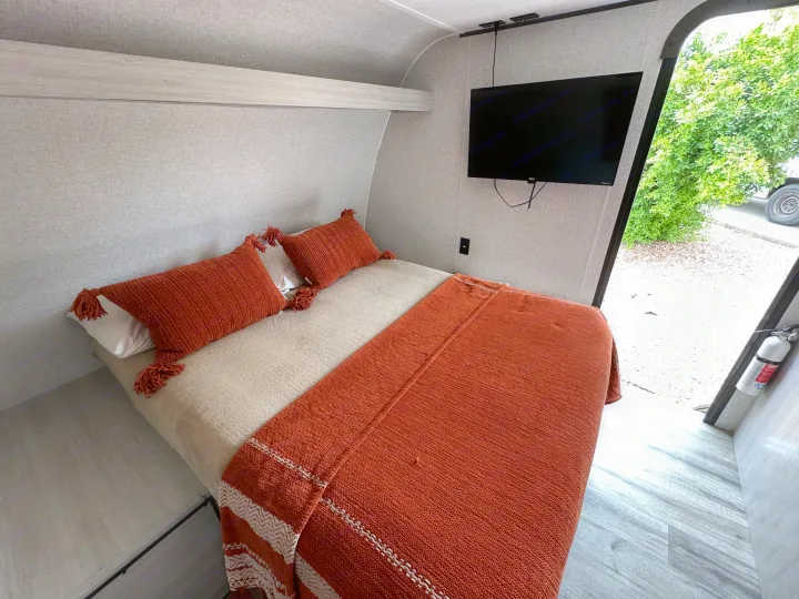 Experience comfort in this RV rental bedroom: a cozy bed with orange-tasseled pillows and a blanket, plus a wall-mounted TV.. Crossroads RV Zinger Lite 2026