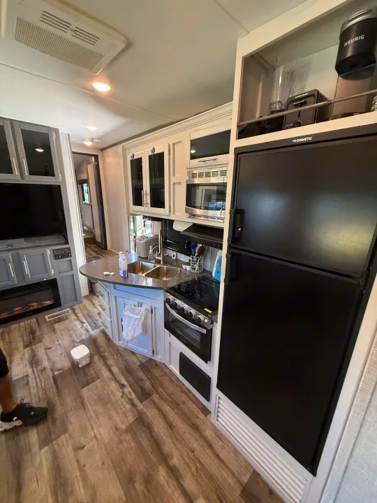 Bright RV rental interior: kitchen with stove, fridge, microwave, double sink, plus living area with TV & fireplace.. Crossroads RV Volante 2022