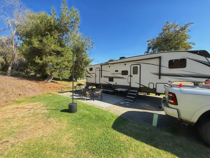 White Volante RV rental at a sunny campsite with a truck, chairs, and grill. Green and dry grass, trees in the background.. Crossroads RV Volante 2022